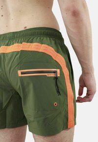 Pantaloni da mare verdi con strisce laterali arancioni, tasca con zip sul retro, vita elastica e pratico dettaglio con cordino.