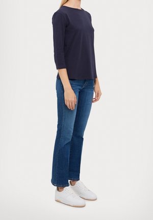 Femme portant un haut bleu marine à manches trois-quarts, un jean droit délavé moyen et des baskets blanches, debout devant un fond uni.