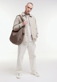 Giacca esterna beige con bottoni a pressione, t-shirt grigio chiaro, pantaloni bianchi e scarpe da ginnastica color crema. Borsa a spalla oversized marrone con dettaglio a fiocco.