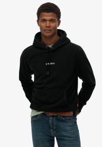 Sudadera de forro polar negra con cordones, bolsillo frontal y logo blanco de "SUPERDRY". Llevada sobre una camiseta verde clara, combinada con jeans azules.