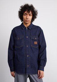 Denim jacket in dark blue, featuring a button-front closure, two chest pockets, and a branded leather patch on the left side.