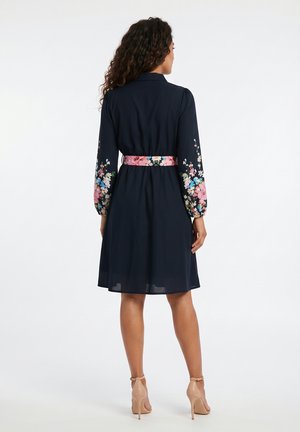 Femme en robe bleu marine avec broderie florale sur les manches et une ceinture fleurie, portant des escarpins à bride à la cheville couleur nude, vue de dos.