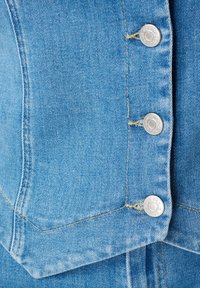 Close-up van een lichtblauwe denimjack, met vier zilveren knoppen aan de voorkant en zichtbare stiksels langs de naden.