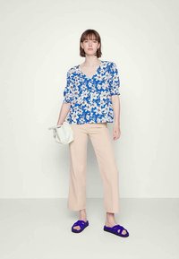 Mulher em pé contra um fundo simples, vestindo calças bege, uma blusa floral azul, sandálias roxas e segurando uma carteira branca.