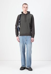 Dark green hoodie with drawstrings, paired with light blue jeans. Model wears a black crossbody bag and black shoes. Solid color, relaxed fit.