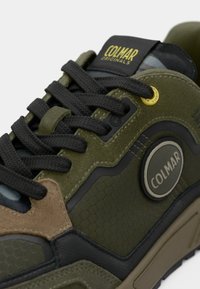 Adidași din suede verde și textil, cu șireturi negre, modele hexagonale și un ecuson logo circular. Accente în nuanțe de bej și negru.