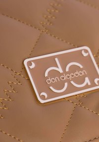 Bolso de mano de imitación cuero marrón con acolchado, que presenta costuras doradas y una placa rectangular con el nombre de la marca en blanco.