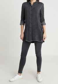 Donna che indossa una tunica nera a maniche lunghe con bottoni, jeans skinny grigi scuri e sneakers bianche, in piedi davanti a uno sfondo neutro.