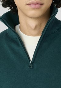 Pull en maille vert foncé avec col zippé. Présente une texture côtelée et une couche intérieure crème contrastante visible au niveau du cou.