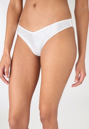 Donna che indossa slip bikini bianchi a vita bassa decorati con piccoli borchie argentee uniformemente distanziate su uno sfondo chiaro e uniforme.