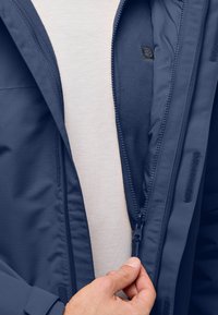 Giacca blu navy con chiusura a zip, con fodera in morbido pile. Presenta un tessuto esterno testurizzato e un'etichetta nera vicino alla zip.