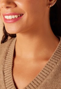 Pull en laine beige avec un décolleté en V. Teint de peau lisse et une subtile boucle d'oreille dorée. Sourire éclatant avec des dents blanches visibles.