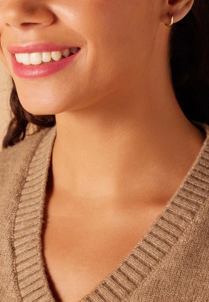 Beige gebreide trui met een V-hals. Glad huidskleur en een subtiele gouden ringoorbel als accent. Heldere glimlach met zichtbare witte tanden.