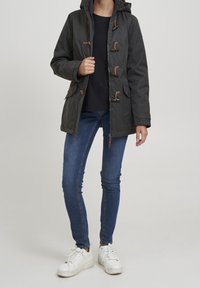 Person som bär en mörkgrå huva med knäppning, blå smala jeans, svart tröja och vita sneakers som står mot en ljus bakgrund.