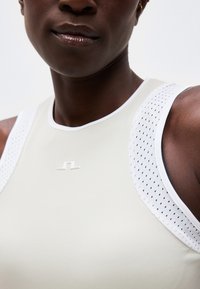 Κοντινό πλάνο ενός ατόμου που φοράει λευκό αθλητικό τοπ χωρίς μανίκια με διάτρητα πάνελ στους ώμους και μικρό λογότυπο στο στήθος.