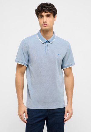 Junger Mann steht und trägt ein hellblaues Poloshirt mit kurzen Ärmeln und weißer Einfassung, kombiniert mit dunkelblauen Jeans, vor einem einfarbigen Hintergrund.