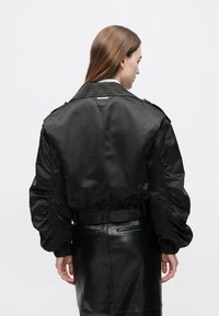 Čierna bomber bunda s lesklou, hladkou textúrou, nariasenými rukávmi a stiahnutým pásom, vybavená elegantným golierom a minimálnymi kovovými doplnkami.