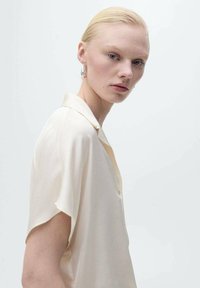 Cream-colored, short-sleeve shirt with a soft sheen, featuring a collar and front buttons. The fabric has a lightweight, smooth texture.
