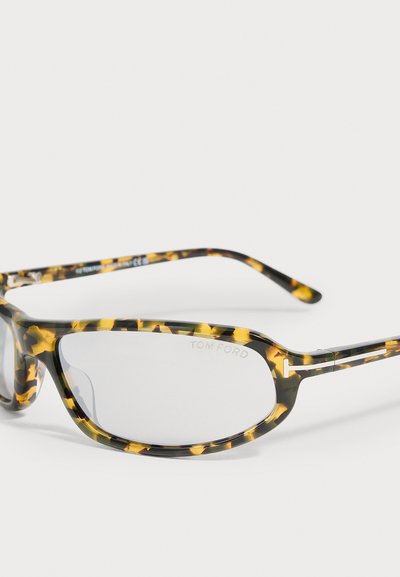 Lunettes avec montures ovales en écaille de tortue et verres transparents, posées en biais sur une surface blanche, avec le logo Tom Ford sur le verre droit.
