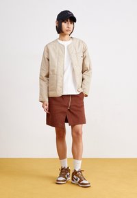 Beige quilted bomber jacket, white T-shirt, brown denim skirt with front zipper, white socks, brown and gray sneakers, black cap.