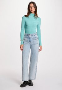 Un pull col roulé à côtes bleu clair, associé à un jean flare en denim clair taille haute et des bottes noires à semelle épaisse. Deux poches plaquées à l'avant sont visibles.