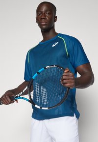 Blå idretts skjorte med mesh tekstur og grønne detaljer, parret med en svart tennisracket med et blått grep. Hvite shorts fullfører antrekket.
