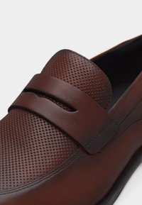 Καφέ μοκασίνι από δέρμα με υφή διάτρητου πάνω μέρους και λείο λουράκι στο πρόσθιο τμήμα, απεικονίζεται πάνω σε απλό λευκό φόντο.