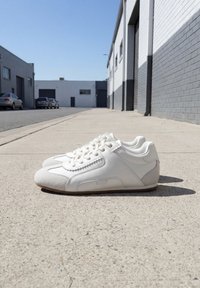 Paire de baskets blanches décontractées posées sur un trottoir en béton à côté d'une rue avec des bâtiments industriels sous un ciel bleu clair.