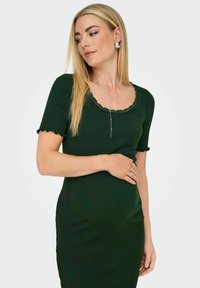 Femme blonde portant une robe côtelée vert foncé avec un col en dentelle, un collier et des boucles d'oreilles argentés, tenant doucement son abdomen et regardant sur le côté.