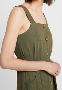 Abito verde oliva con scollatura dritta, spalline larghe, grandi bottoni e tasche laterali. Realizzato in un tessuto leggero e strutturato.