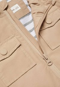 Ljusbeige jacka med en slät, matt finish. Har en synlig dragkedja, två framfickor och ett randigt tygfoder.