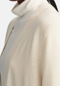 Krämfärgad cardigan med en lätt stickad textur, öppen framsidan och subtila ribbade detaljer i kanterna. Turtleneck under.