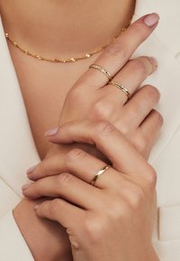 Goldene Ringe mit unterschiedlichen Designs, darunter gedrehte, geflochtene und facettierte Muster, getragen an leicht manikürten Händen. Eine feine goldene Kettenuhr ist sichtbar.