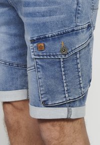 Blå denim cargo shorts med opfoldede kanter, flere lommer, tan læderlap og mørke hardware detaljer. Båret på et ben med lyst hår.