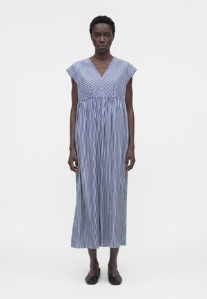 Žena nesúca modro-bielo pruhované midi šaty bez rukávov s výstrihom do V a čierne baleríny, stojaca na jednoduchom bielom pozadí.