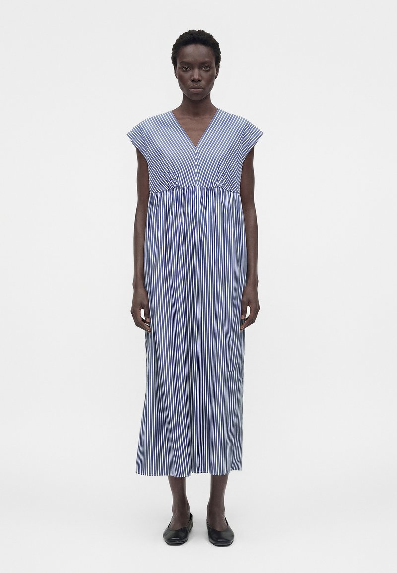 Γυναίκα που φοράει μπλε και άσπρο κάθετα ριγέ, αμάνικο midi φόρεμα με V-λαιμόκοψη και μαύρες μπαλαρίνες, στέκεται μπροστά σε ένα απλό λευκό φόντο.