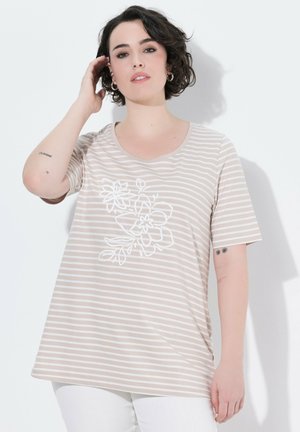Donna con capelli corti e scuri che indossa una maglietta a righe beige e bianca con stampa floreale bianca e pantaloni bianchi, toccandosi i capelli.
