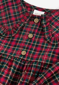 Chemise à carreaux rouge, vert et bleu marine avec des boutons en bois et un col à volants.