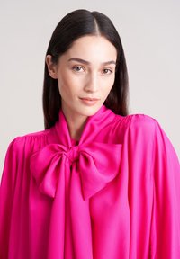 Blusa di un brillante colore rosa con un grande fiocco al collo, realizzata in tessuto liscio. Presenta maniche arricciate e un design fluido.
