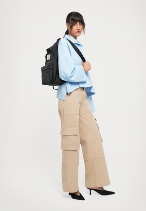 Frau trägt ein hellblaues, übergroßes Hemd, beigen weiten Cargo-Hosen, schwarze Slingback-Absätze und hat einen schwarzen Rucksack über eine Schulter gehängt.