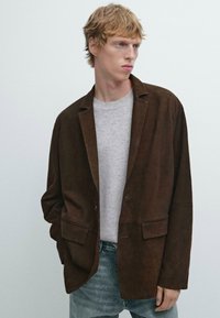 Bruine suède blazer met een losse pasvorm, voorzien van gelapte revers en twee voorzakken, gelaagd over een lichtgrijze trui en denim.