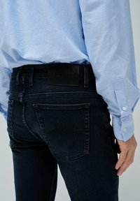 Vaqueros de mezclilla de un azul oscuro con un parche de cuero de Salsa Jeans en la parte posterior, combinados con una camisa de manga larga de un azul claro. Ajuste entallado y textura suave.
