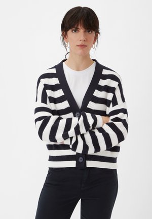 Gestreifter Cardigan in Marineblau und Weiß, gestrickte Textur, V-Ausschnitt, vorn mit Knopfleiste, und cropped Schnitt. Kombiniert mit einem weißen Oberteil und schwarzen Hosen.