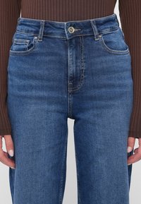 Jeans de denim azul de cintura alta con un diseño clásico de cinco bolsillos, herrajes de botón de cobre y un leve desvanecimiento a lo largo de los muslos.