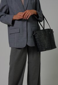 Una persona indossa un completo grigio con pantaloni a gamba larga e tiene in mano una borsa a tracolla nera con un motivo su uno sfondo grigio uniforme.