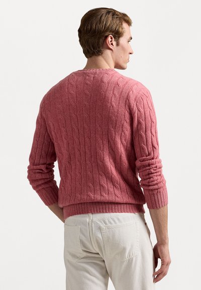 Suéter de punto cableado en rosa empolvado, con puños y dobladillo de canalé, textura de peso medio, presenta un patrón sutil, combinado con pantalones de color claro.