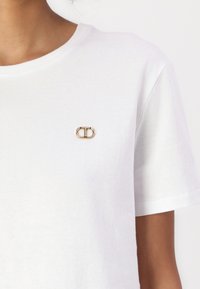 T-shirt bianco in cotone con collo rotondo, con un logo in oro e cristallo sul lato sinistro del petto. Maniche corte, texture morbida.