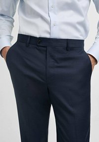 Pantalons de costume bleu marine avec un devant plat, passants de ceinture, un bouton unique et deux poches latérales. Le matériau semble lisse et légèrement texturé.