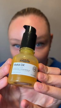 Personne tenant une bouteille d'huile concentrée professionnelle L'Oréal Metal DX près de la caméra, avec des yeux bleus visibles en arrière-plan.