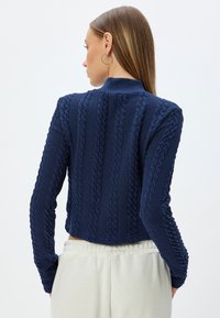 Mörkblå kabelstickad cardigan med en kort design och ribbad krage. Har texturerade mönster längs ärmarna och bakstycket.